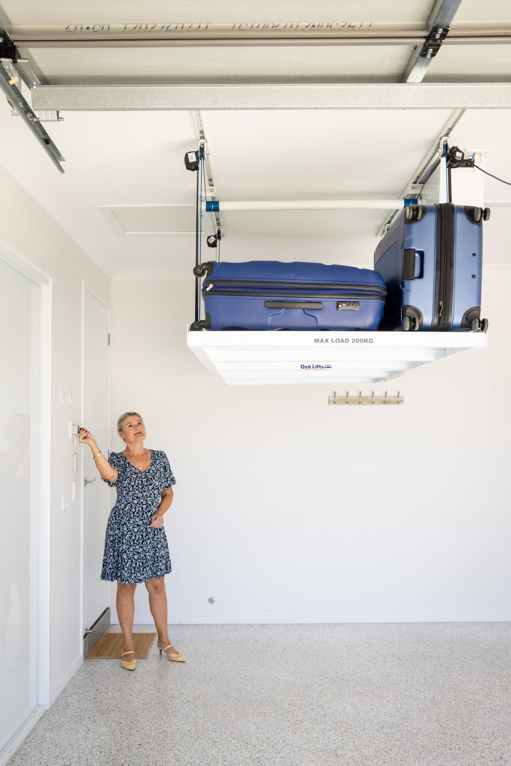 Woman in a blue floral dress reaching to operate a ceiling-mounted luggage hoist carrying two blue suitcases in a bright room.